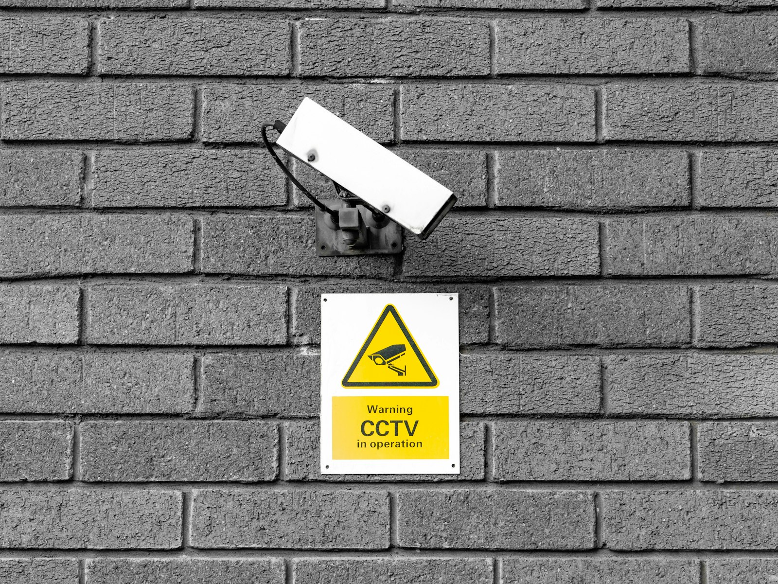 Videonovērošanas kamera pie ķieģeļu sienas ar dzeltenu CCTV brīdinājuma zīmi
